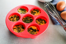 Load image into Gallery viewer, Red silicone egg bites mould with egg mixture inside on a worktop with whisk and eggs