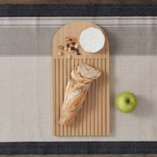 Load image into Gallery viewer, A beige bamboo arch bread board with grooves for catching crumbs, with bread and nuts and cheese on the board. the board is on a striped tablecloth and an apple it next to the board