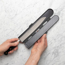Load image into Gallery viewer, Knife with a black handle and gray sheath held by a hand on a light gray marble background