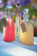 Load image into Gallery viewer, Three clear glass bottles with handwritten labels and coloured liquid inside on a pale table with a blurred floral background.