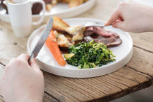 Load image into Gallery viewer, A person is using a white ceramic dinner plate with a subtle swirl pattern inside, which is set on a wooden table with a meal consisting of roasted vegetables.