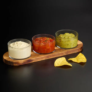 Three glass bowls with different dips on a wooden board with tortilla chips on a black background