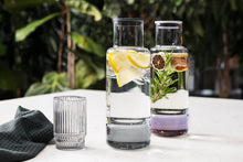 Load image into Gallery viewer, Two glass carafes with water and lemon, lime, and rosemary garnishes on a white surface with a blurred green outdoor background.
