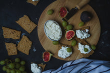 Load image into Gallery viewer, Wooden board with crackers, figs, grapes, and a bowl of cream cheese on a dark surface.
