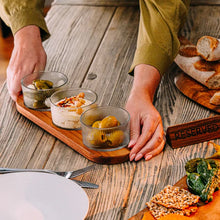 Load image into Gallery viewer, Person arranging small glass bowls with snacks on a wooden tray, with bread and other food items on a table.
